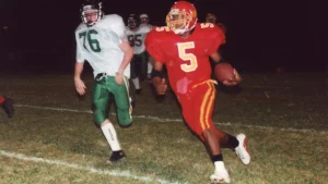 A high school running back in a red and gold uniform carries the ball upfield at night as a defender in white and green gives chase.