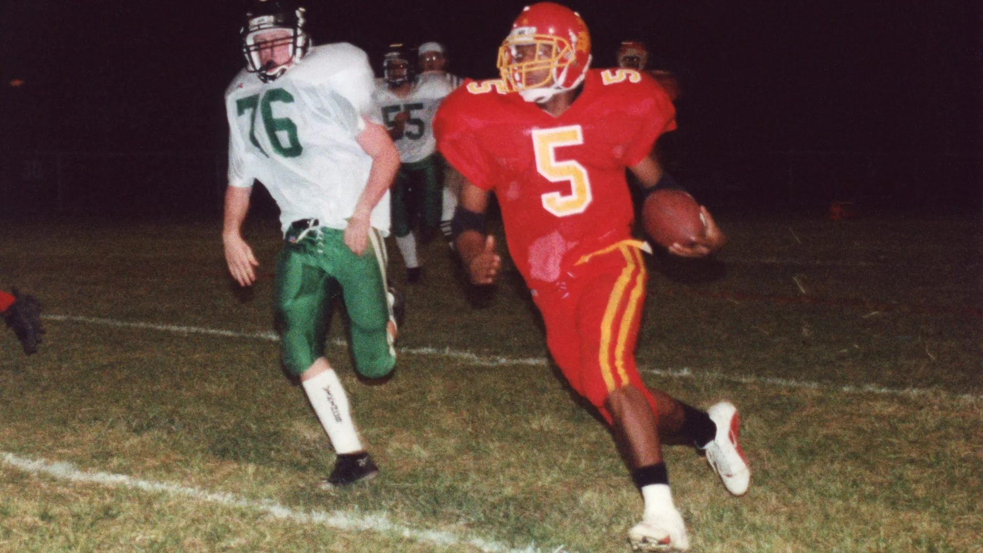 A high school running back in a red and gold uniform carries the ball upfield at night as a defender in white and green gives chase.
