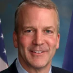 Official portrait of U.S. Sen. Dan Sullivan of Alaska in a dark suit, with the U.S. flag and the Alaska state flag behind him.