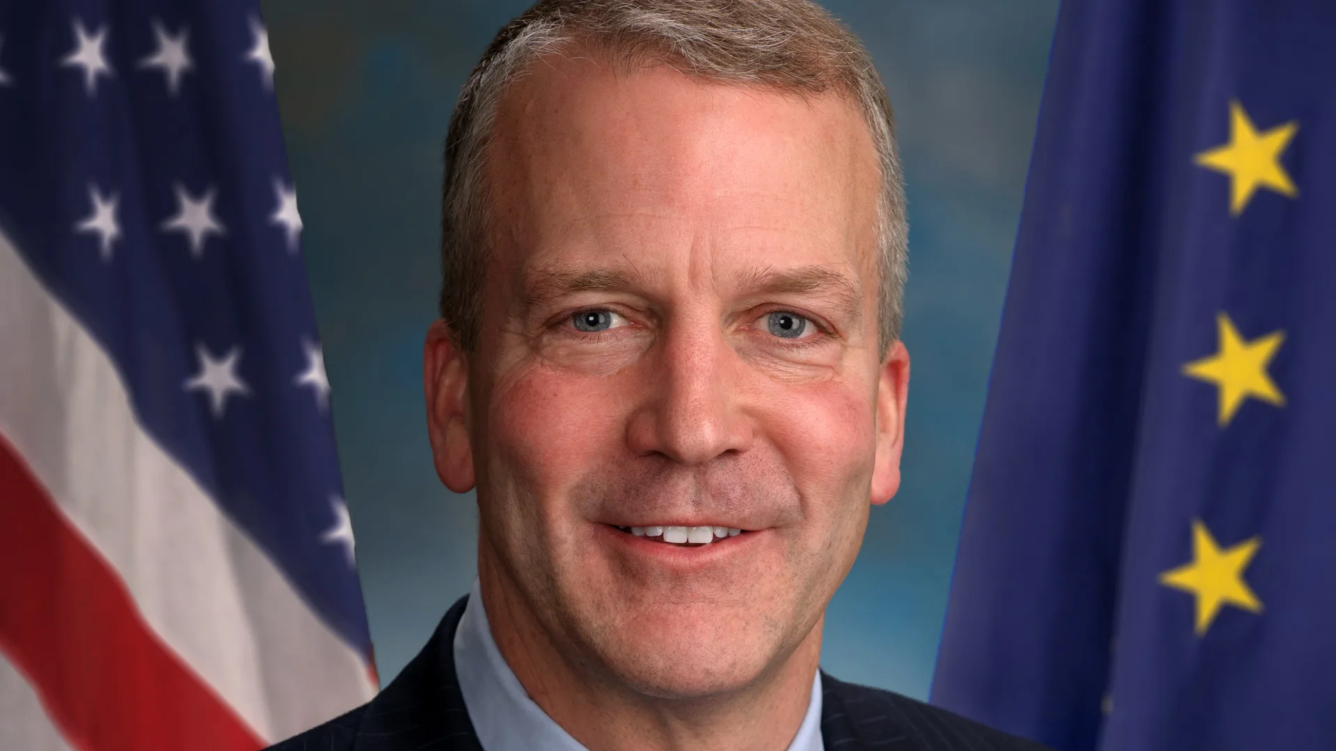 Official portrait of U.S. Sen. Dan Sullivan of Alaska in a dark suit, with the U.S. flag and the Alaska state flag behind him.
