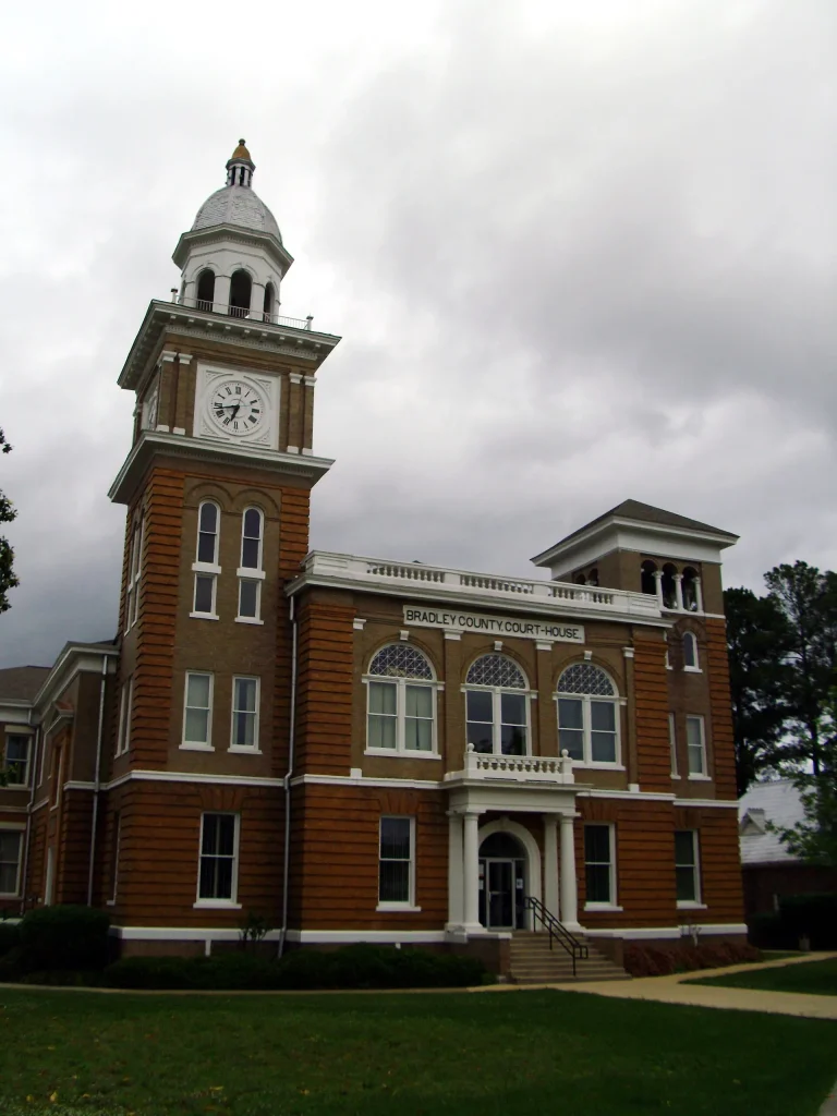 Two-story red brick Bradley County Courthouse with white trim, a columned entrance porch and a tall clock tower topped by a white cupola, under a cloudy sky.