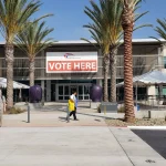 A "Vote Here" banner hangs on the front of the San Diego County Registrar of Voters building, with palm trees and voters on the walkway, during early voting in October 2018.