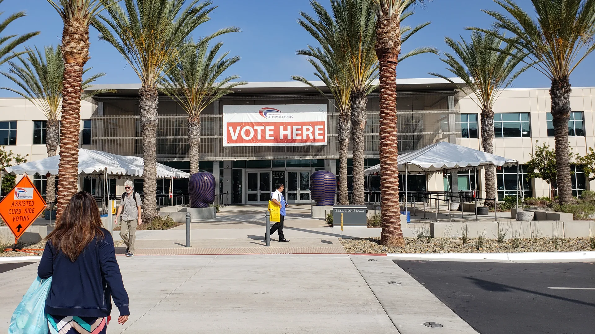 A "Vote Here" banner hangs on the front of the San Diego County Registrar of Voters building, with palm trees and voters on the walkway, during early voting in October 2018.