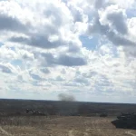 Armored vehicles spread across an open, dry training range under a cloudy sky as smoke rises in the distance at Wainwright, Alberta