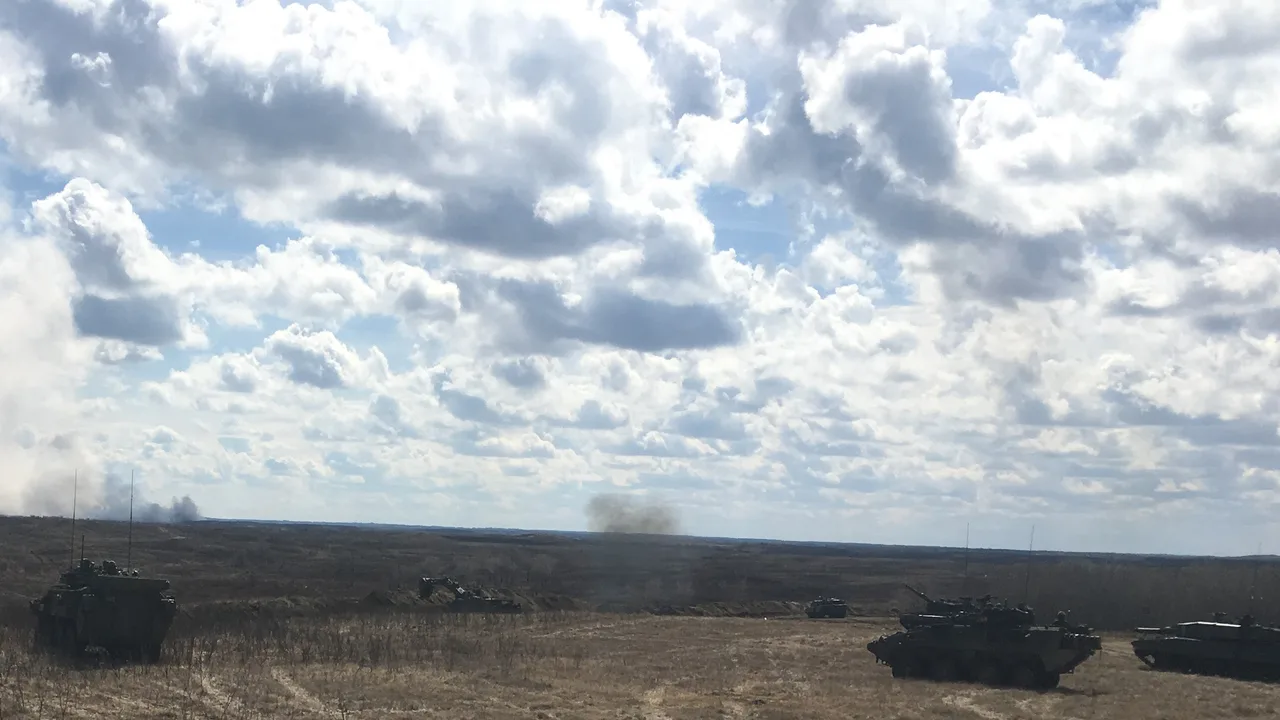Armored vehicles spread across an open, dry training range under a cloudy sky as smoke rises in the distance at Wainwright, Alberta