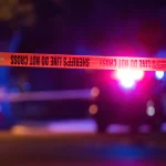 Yellow sheriff's line tape reading "Do Not Cross" stretched across a street at night, with the flashing lights of a patrol car blurred in the background.