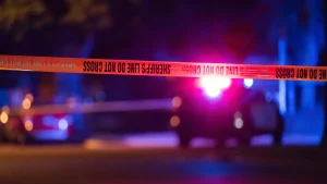 Yellow sheriff's line tape reading "Do Not Cross" stretched across a street at night, with the flashing lights of a patrol car blurred in the background.