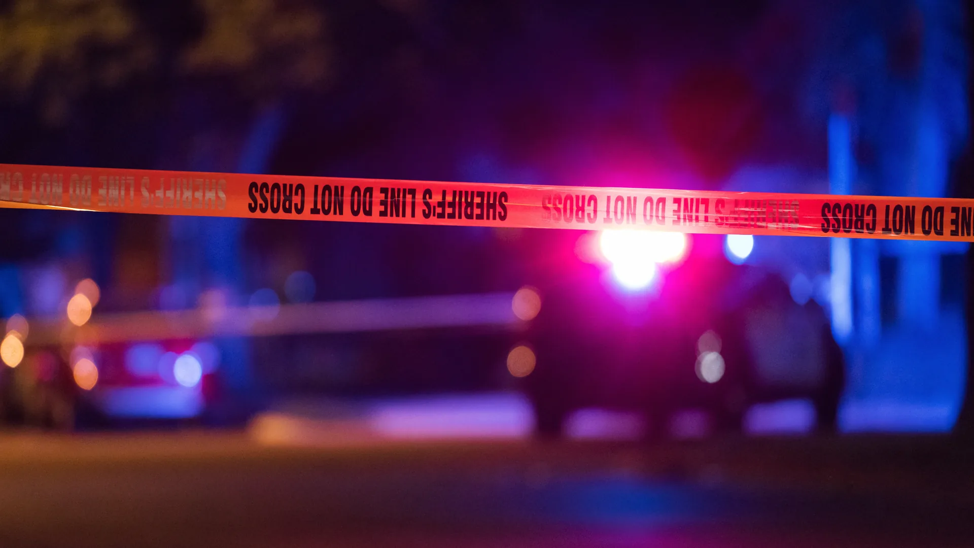 Yellow sheriff's line tape reading "Do Not Cross" stretched across a street at night, with the flashing lights of a patrol car blurred in the background.