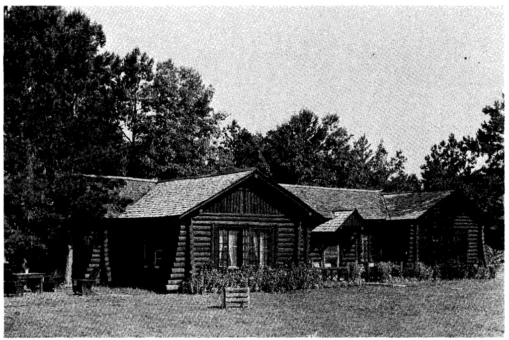 Black-and-white photo of a long one-story log house with a shingled gable roof and a smaller gabled porch, set on a lawn in front of tall pines and hardwoods.