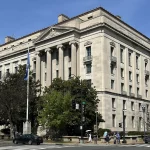 The limestone Robert F. Kennedy Department of Justice Building, with tall columns and a flag out front, on a corner in Washington, D.C., under a clear blue sky.