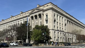 The limestone Robert F. Kennedy Department of Justice Building, with tall columns and a flag out front, on a corner in Washington, D.C., under a clear blue sky.