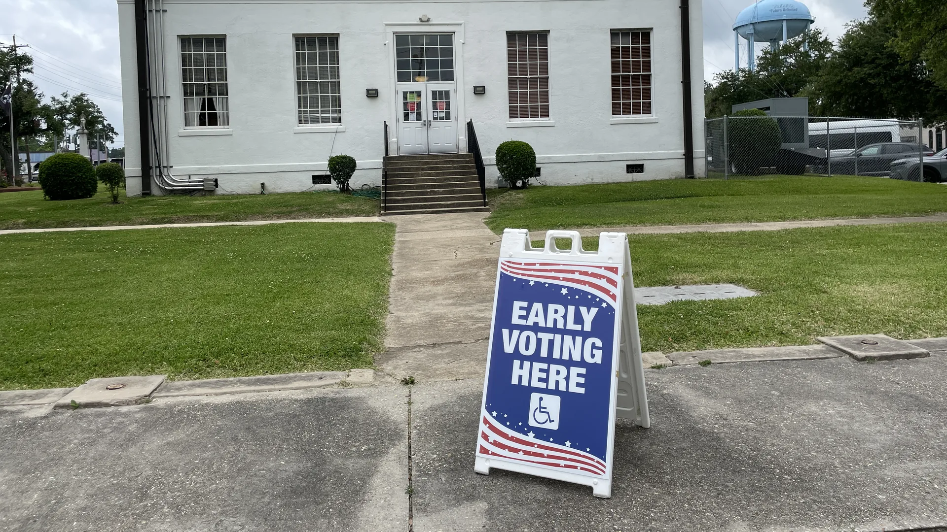 A blue Early Voting Here sandwich-board sign on a sidewalk in front of a white government building