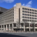 The tan concrete J. Edgar Hoover Building, headquarters of the FBI, seen from across Pennsylvania Avenue in Washington under a blue sky.