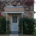 The small tan brick Hermitage City Hall and Jail building in Hermitage, Arkansas, with a white door, a shingled awning and shrubs out front.