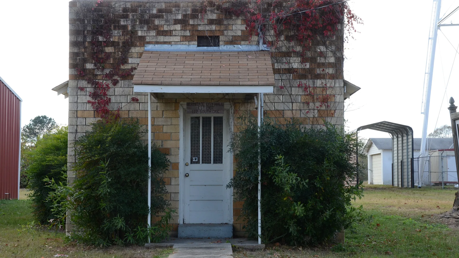 The small tan brick Hermitage City Hall and Jail building in Hermitage, Arkansas, with a white door, a shingled awning and shrubs out front.