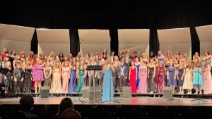 A large high school choir in formal wear performs on a stage as audience members watch from theater seats