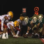 Two high school football teams, one in white and red and one in green and gold, crouched at the line of scrimmage before the snap at a night game.
