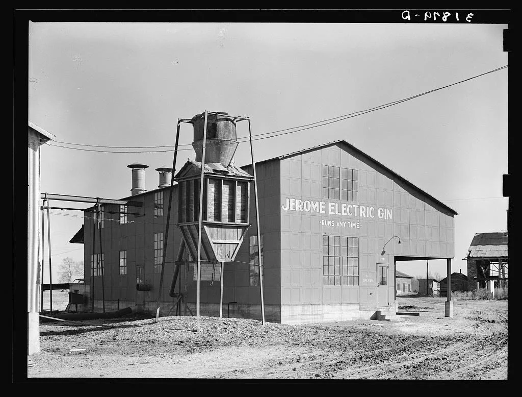 A metal-sided cotton gin building painted with the words Jerome Electric Gin, with a cyclone separator tower beside it.
