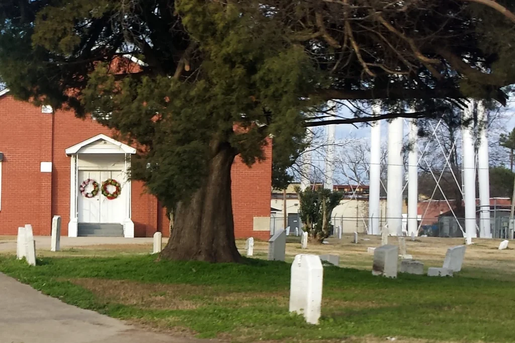 White grave markers under a large tree in the foreground, with a small white church entrance decorated with wreaths beside a red brick building behind.