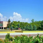 Flower beds and trees along the central square in Oleshky, Ukraine, with a pink two-story building and a monument in the background under a blue sky.