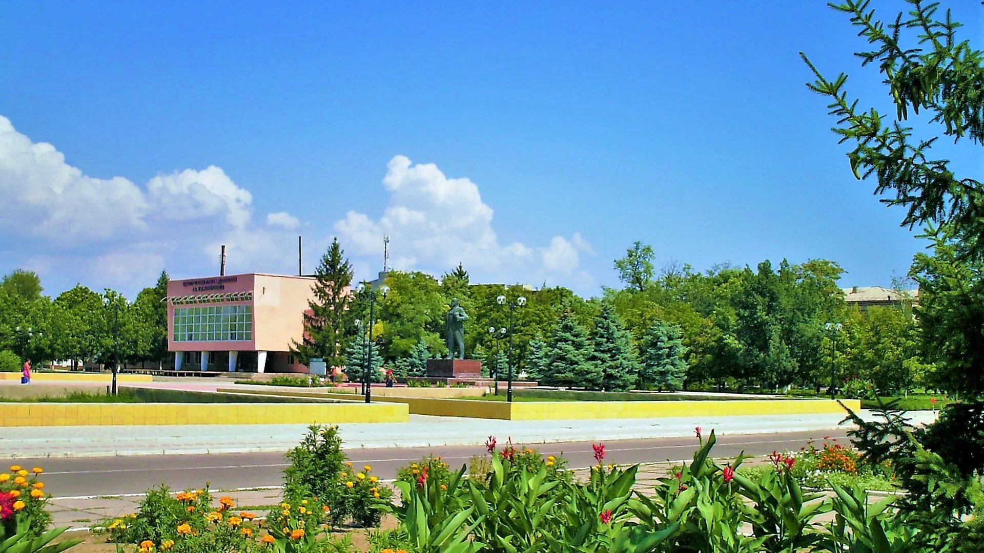 Flower beds and trees along the central square in Oleshky, Ukraine, with a pink two-story building and a monument in the background under a blue sky.