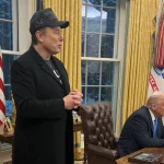 Elon Musk, in a black coat and cap, stands and talks in the Oval Office as President Donald Trump sits at the Resolute Desk in February 2025.