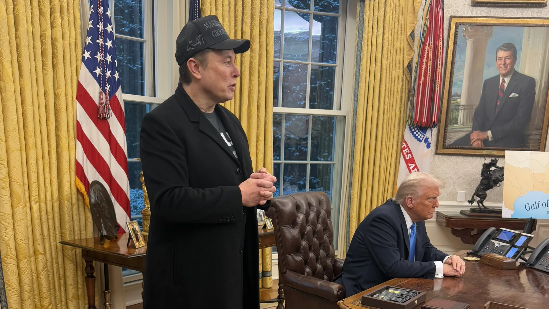 Elon Musk, in a black coat and cap, stands and talks in the Oval Office as President Donald Trump sits at the Resolute Desk in February 2025.
