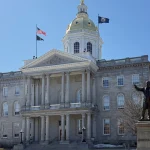 The New Hampshire State House, a granite building with a gold dome and columned portico, in Concord on a clear day.