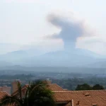 A tall plume of wildfire smoke rises over mountains beyond tile-roofed homes in Orange County, California
