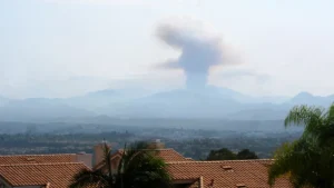A tall plume of wildfire smoke rises over mountains beyond tile-roofed homes in Orange County, California