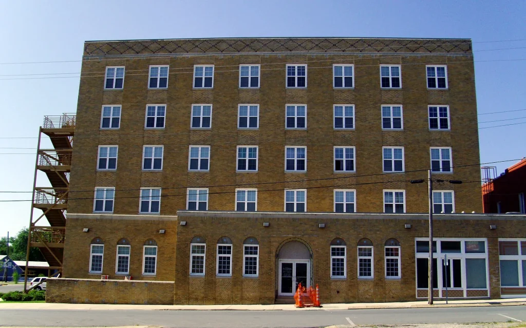 Five-story tan brick building with even rows of windows and a metal fire escape down its left side, on a downtown street corner.