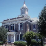 The brick Cleveland County Courthouse in Rison, Arkansas, with a white clock tower, framed by trees under a blue sky.