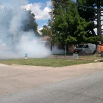 White smoke drifts across a front yard and street from a sewer line during smoke testing in a residential neighborhood in Springdale, Arkansas, as a worker in an orange shirt stands near a utility van.