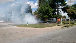 White smoke drifts across a front yard and street from a sewer line during smoke testing in a residential neighborhood in Springdale, Arkansas, as a worker in an orange shirt stands near a utility van.