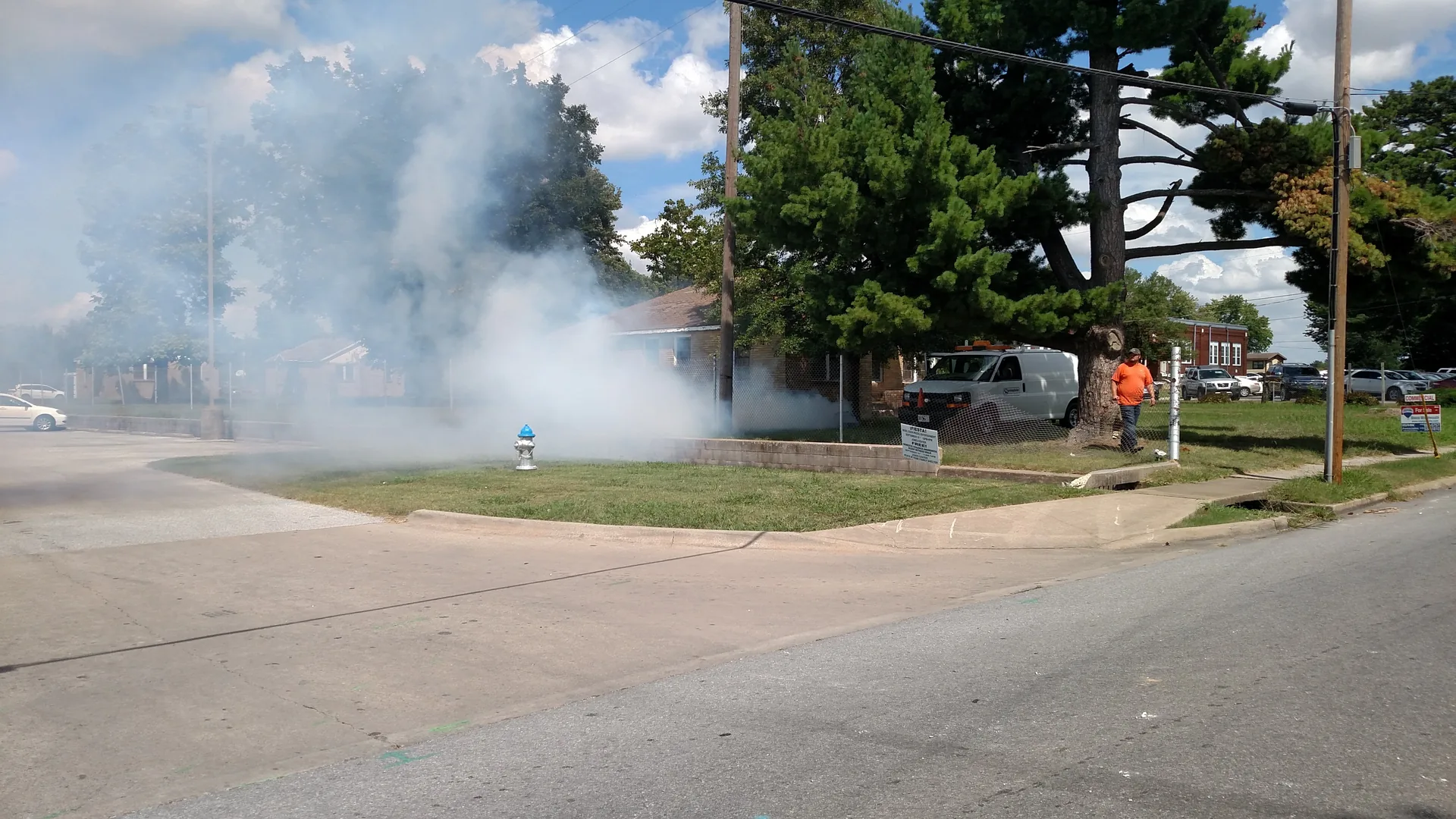White smoke drifts across a front yard and street from a sewer line during smoke testing in a residential neighborhood in Springdale, Arkansas, as a worker in an orange shirt stands near a utility van.