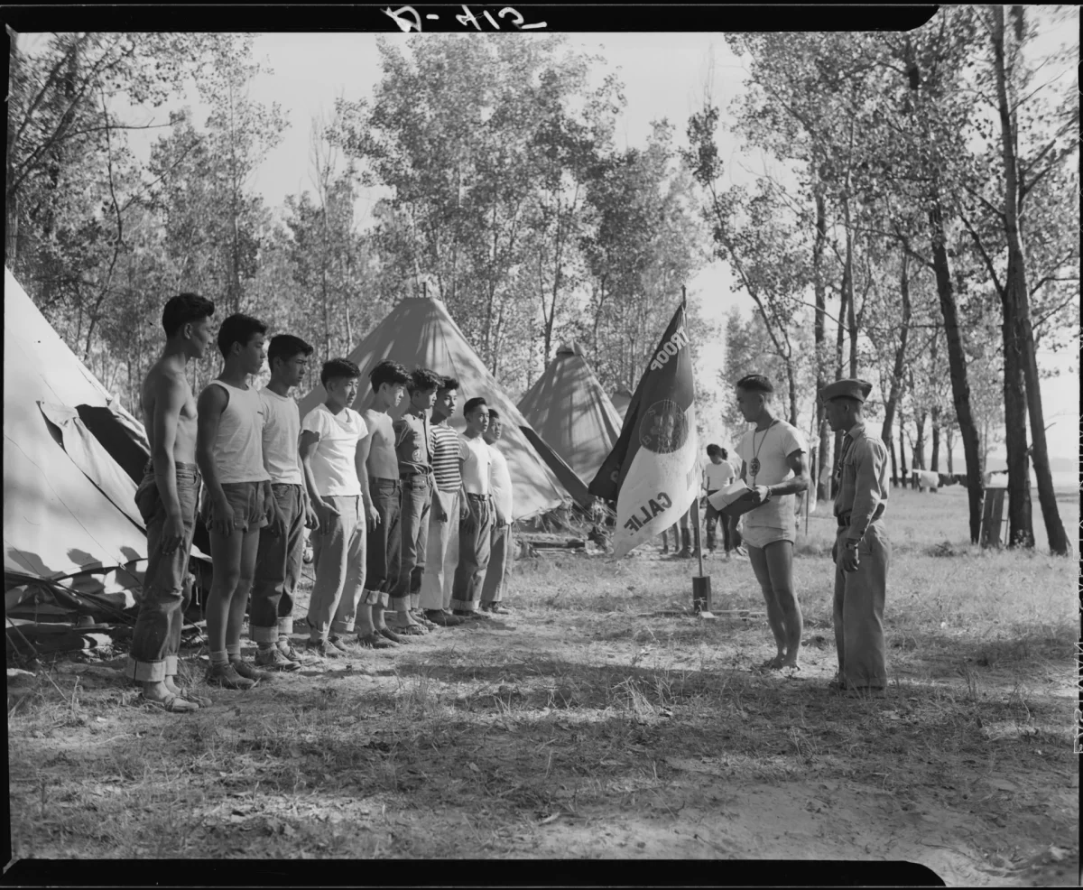 A line of teenage boys stands facing two leaders near canvas tents and a troop flag at a wooded campsite.
