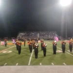 The Warren marching band performs on the field under the stadium lights, with color guard members in the background.