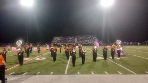 The Warren marching band performs on the field under the stadium lights, with color guard members in the background.