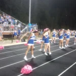 Monticello cheerleaders in blue and white perform on the track in front of the grandstand.