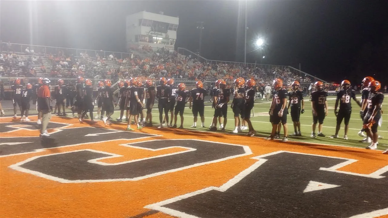 Warren Lumberjacks players in black jerseys and orange helmets stand along the sideline on the orange W at midfield, with the home grandstand behind them under the stadium lights.