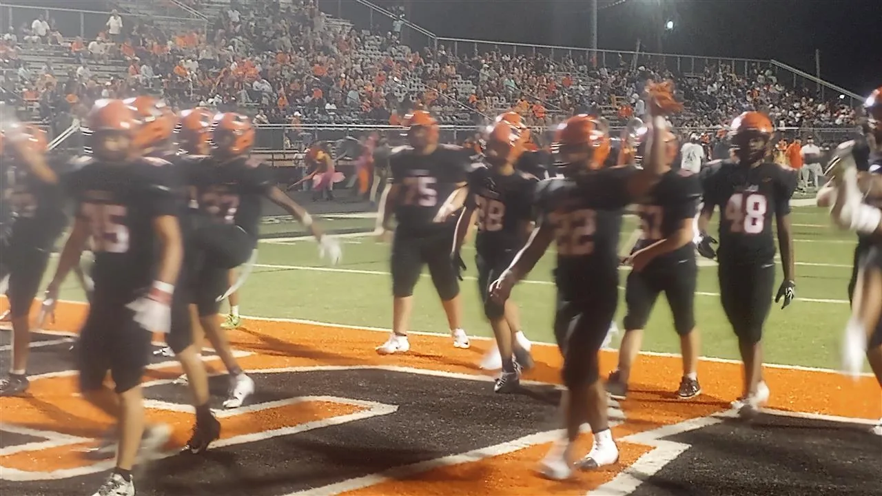 Warren players in black jerseys and orange helmets walk across the field with the grandstand behind them.