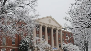 Snow-covered trees frame Building 1 of the National Institutes of Health, a red brick building with white columns and "National Institutes of Health" lettered above the entrance, in Bethesda, Maryland.