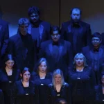 Members of the University of Arkansas at Monticello choir, dressed in black concert attire, sing in rows on a stage under blue lighting.