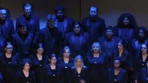 Members of the University of Arkansas at Monticello choir, dressed in black concert attire, sing in rows on a stage under blue lighting.