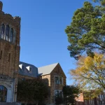 The historic limestone Music Building with its tall stone tower on the University of Arkansas at Monticello campus, with a pine tree and fall foliage under a clear blue sky.