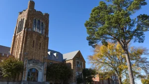The historic limestone Music Building with its tall stone tower on the University of Arkansas at Monticello campus, with a pine tree and fall foliage under a clear blue sky.