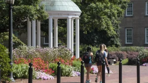 The domed Old Well, a white columned rotunda, on the UNC-Chapel Hill campus, with flower beds and students walking past on a brick path.