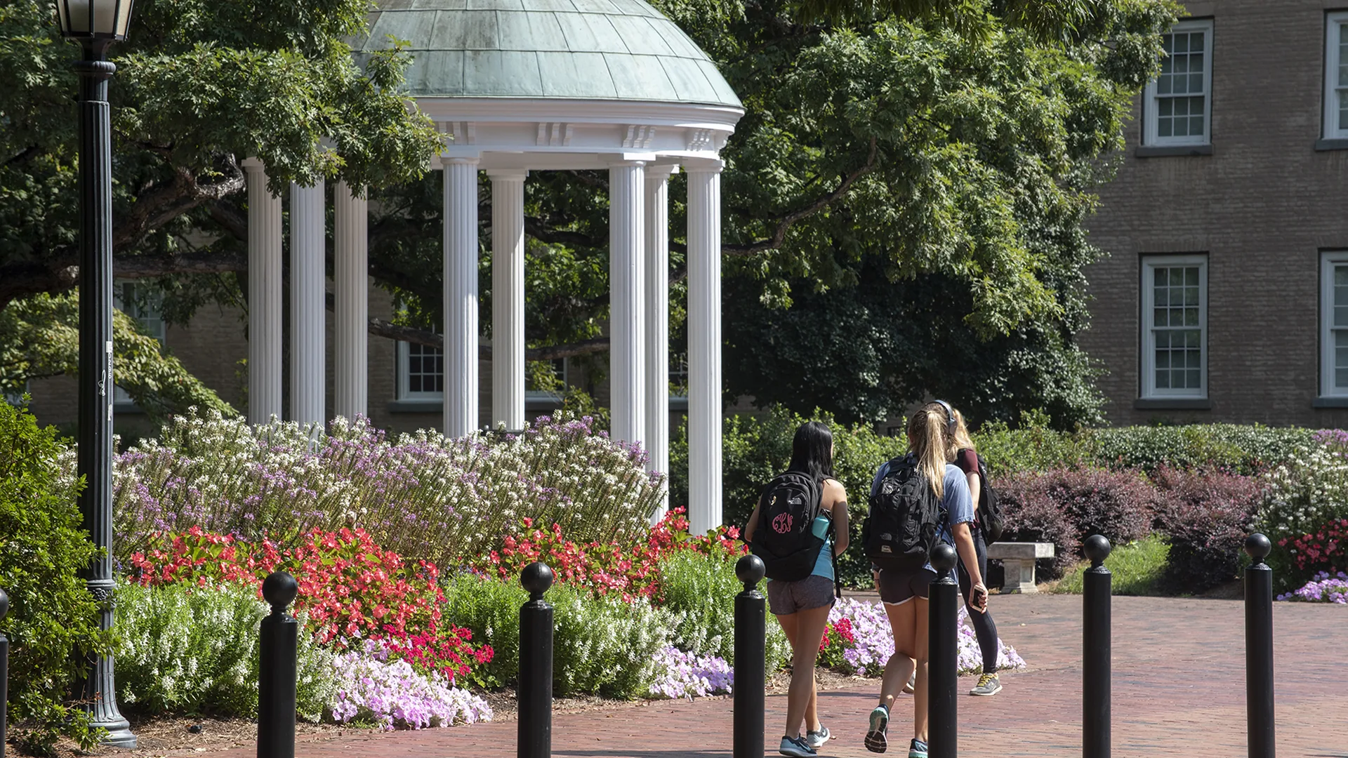 The domed Old Well, a white columned rotunda, on the UNC-Chapel Hill campus, with flower beds and students walking past on a brick path.