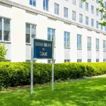 A blue "Department of State" sign on the lawn in front of the Harry S. Truman Building, the State Department headquarters in Washington, D.C., in spring.