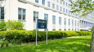 A blue "Department of State" sign on the lawn in front of the Harry S. Truman Building, the State Department headquarters in Washington, D.C., in spring.